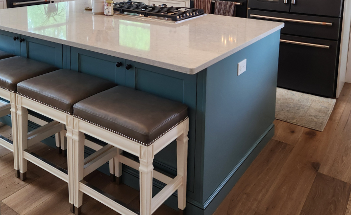 Modern kitchen island with a light countertop, blue cabinet base, and dark upholstered bar stools, with stainless appliances and wood flooring in the background.